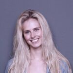 Eine lächelnde Frau mit langen blonden Haaren vor einem grauen Hintergrund, sie strahlt Freundlichkeit und positive Energie aus.