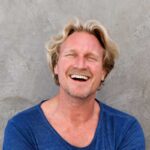 Lächelnder Mann mit blonden, lockigen Haaren und einem blauen Shirt vor einer grauen Wand. Er hat die Arme verschränkt und strahlt Freude aus.