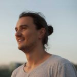 Ein lächelnder Mann mit langen Haaren schaut entspannt in die Ferne, während er in einem Park steht. Der Himmel ist klar und der Abend bricht an.
