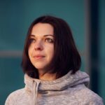 Frau mit kurzen, dunklen Haaren und grauem Pullover schaut nach oben, vor einem blauen Hintergrund. Sanfter Gesichtsausdruck und neugieriger Blick.
