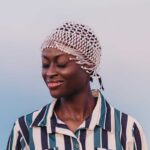 Eine afrikanische Frau mit geschlossen Augen, lächelt sanft. Sie trägt ein gestreiftes Hemd und eine kunstvoll verzierte Kopfbedeckung.