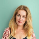 Junge Frau mit langen blonden Haaren lächelt fröhlich vor einer grünen Wand. Sie trägt ein dunkles Oberteil und einen roten, gemusterten Cardigan.